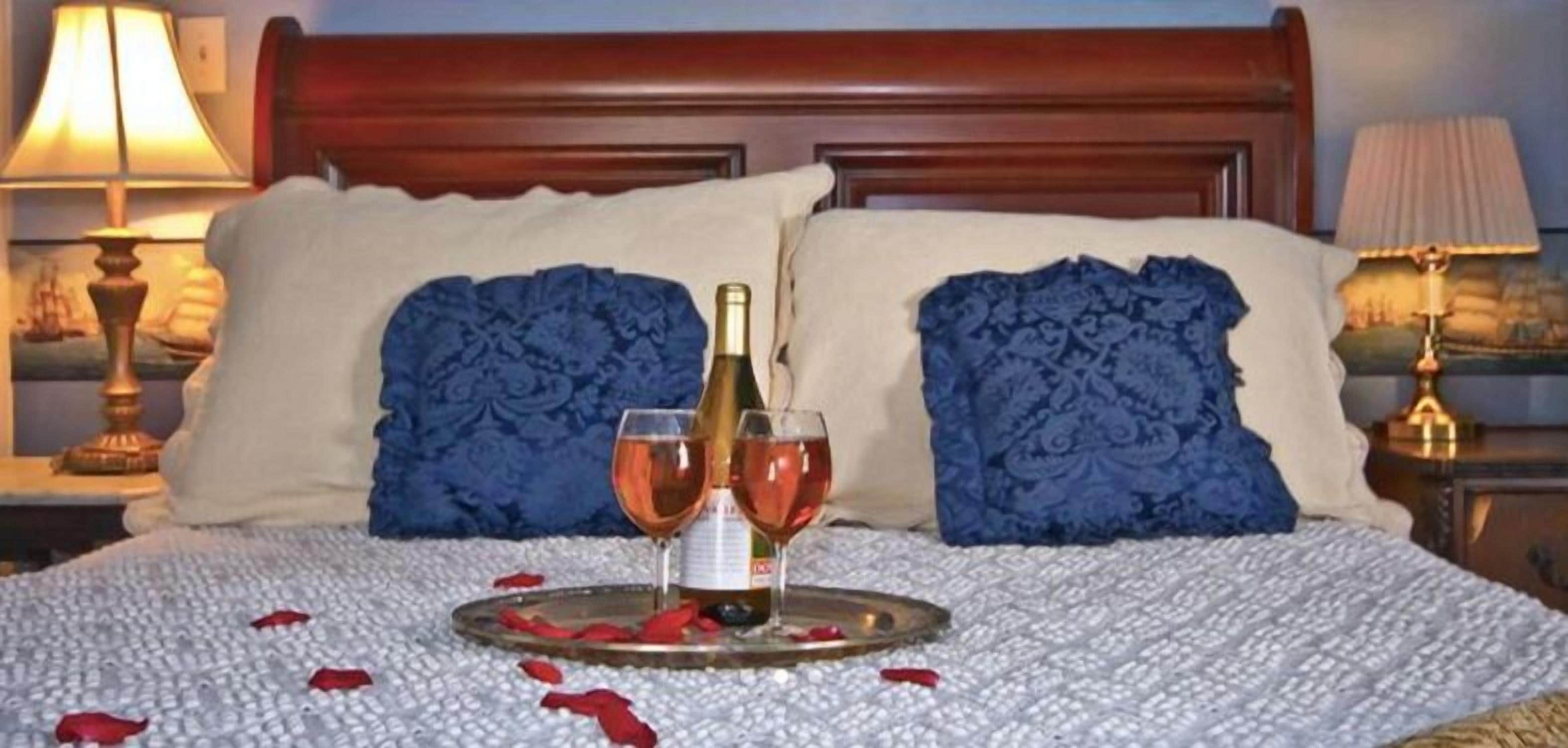 Una cama con una bandeja de pétalos de rosa, dos copas de vino y una botella sobre un edredón blanco y almohadas azules.