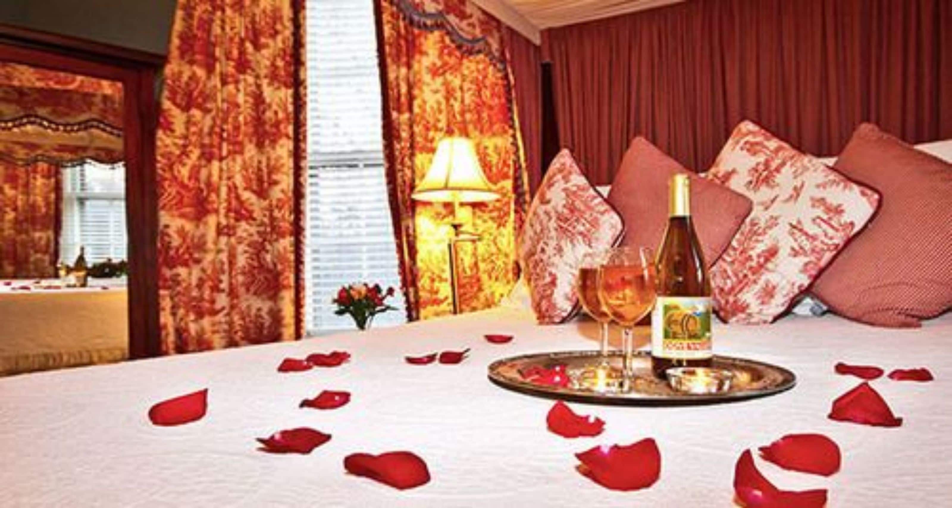 A hotel room with a bed, a bottle of wine, a glass, and rose petals on the bed.