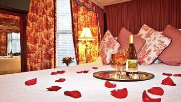 A hotel room with a bed, a bottle of wine, a glass, and rose petals on the bed.
