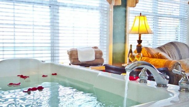 A hotel room with a large bathtub filled with water and rose petals, a sofa, a lamp, and a window with blinds.