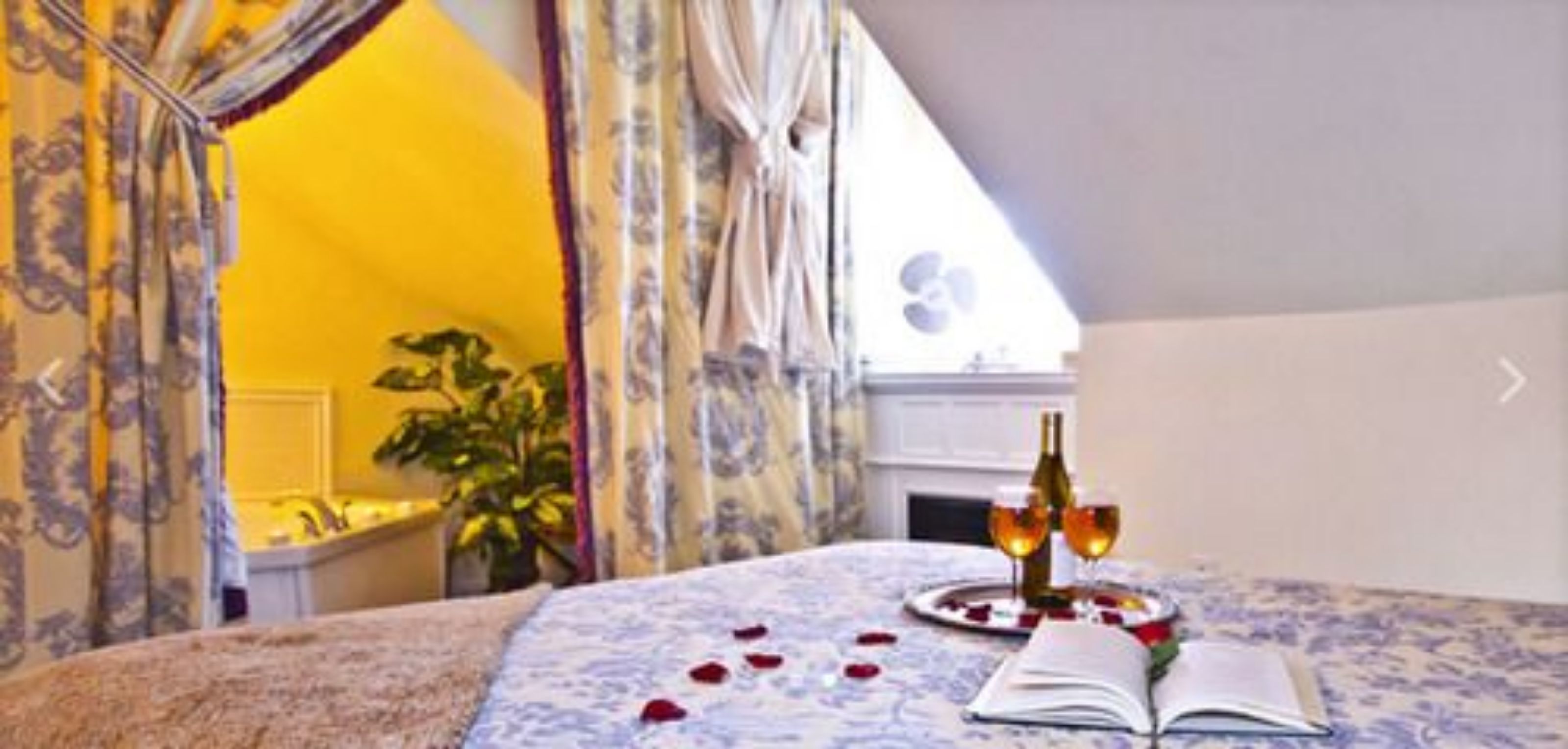 A bedroom with a bed, a bedside table with a bottle and glasses, and a window with curtains.