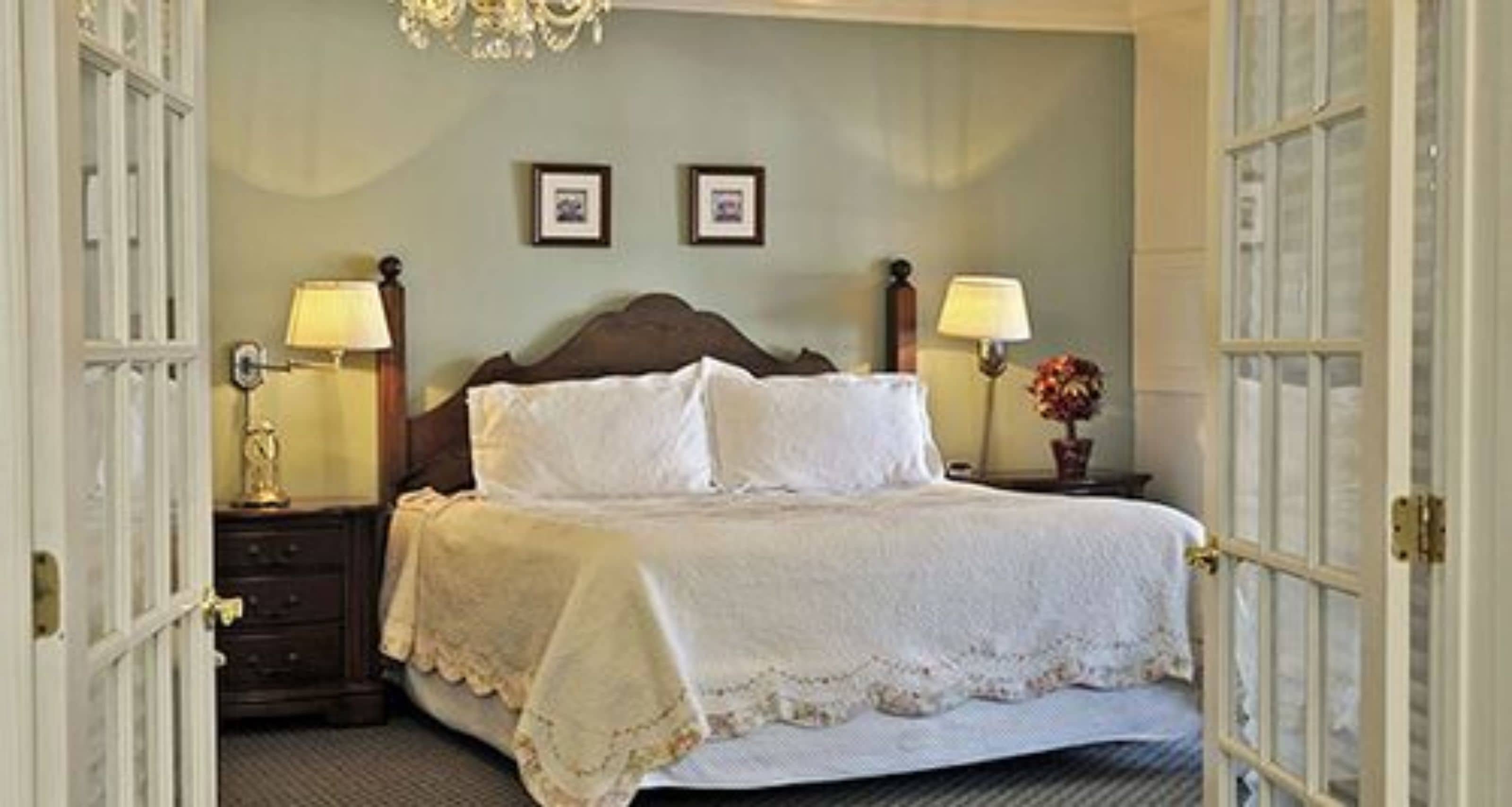 A neatly made bed with a white bedspread and pillows, flanked by two bedside tables with lamps, in a room with a classic design and a chandelier.