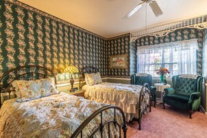 A bedroom with floral wallpaper, two beds, a green armchair, a small table with a vase, and a window with lace curtains.