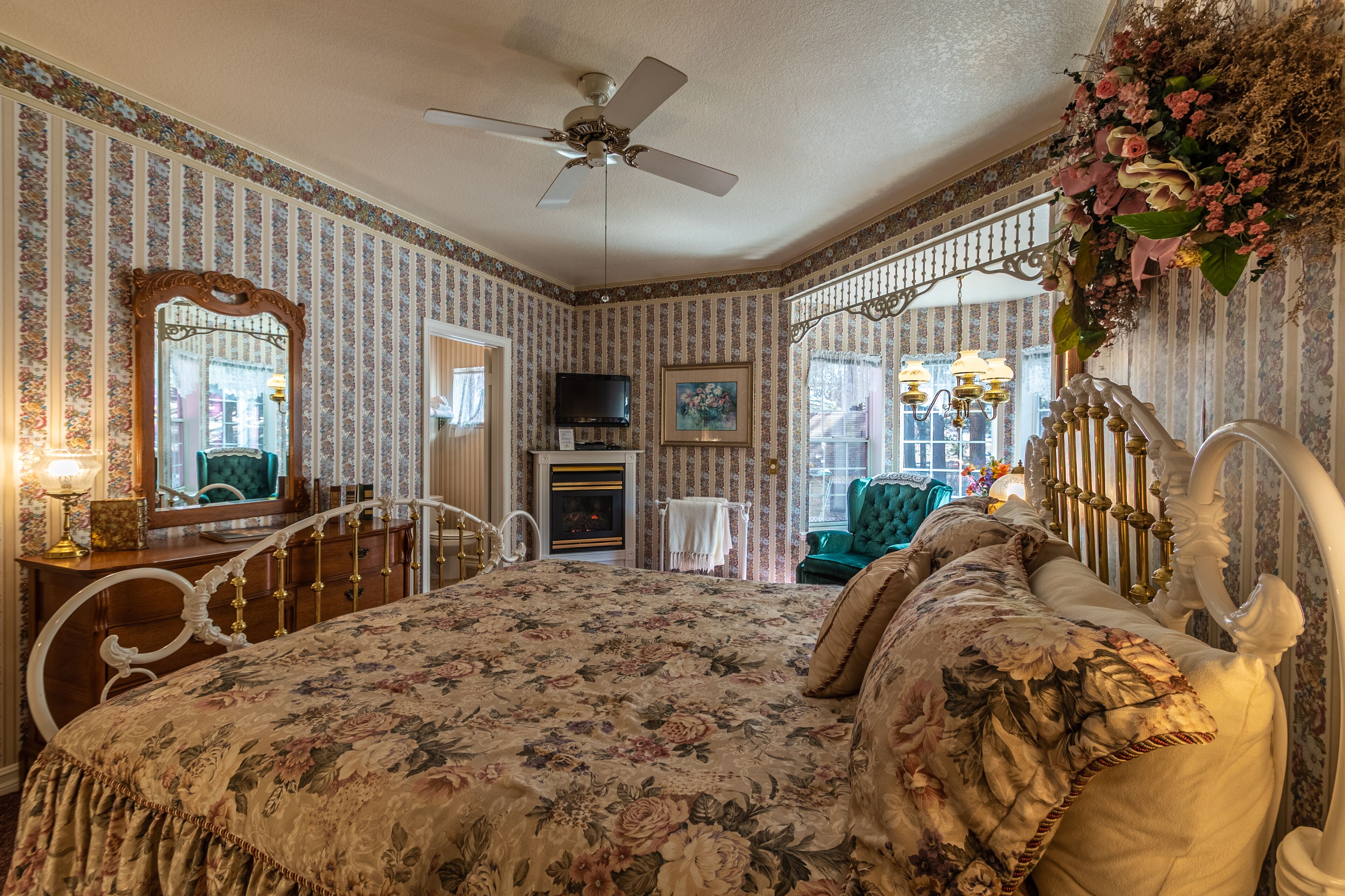 A bedroom with a floral bedspread, a ceiling fan, a fireplace, a television, and a green armchair.
