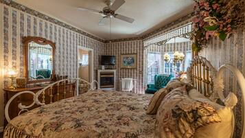 A bedroom with a floral bedspread, a ceiling fan, a fireplace, a television, and a green armchair.