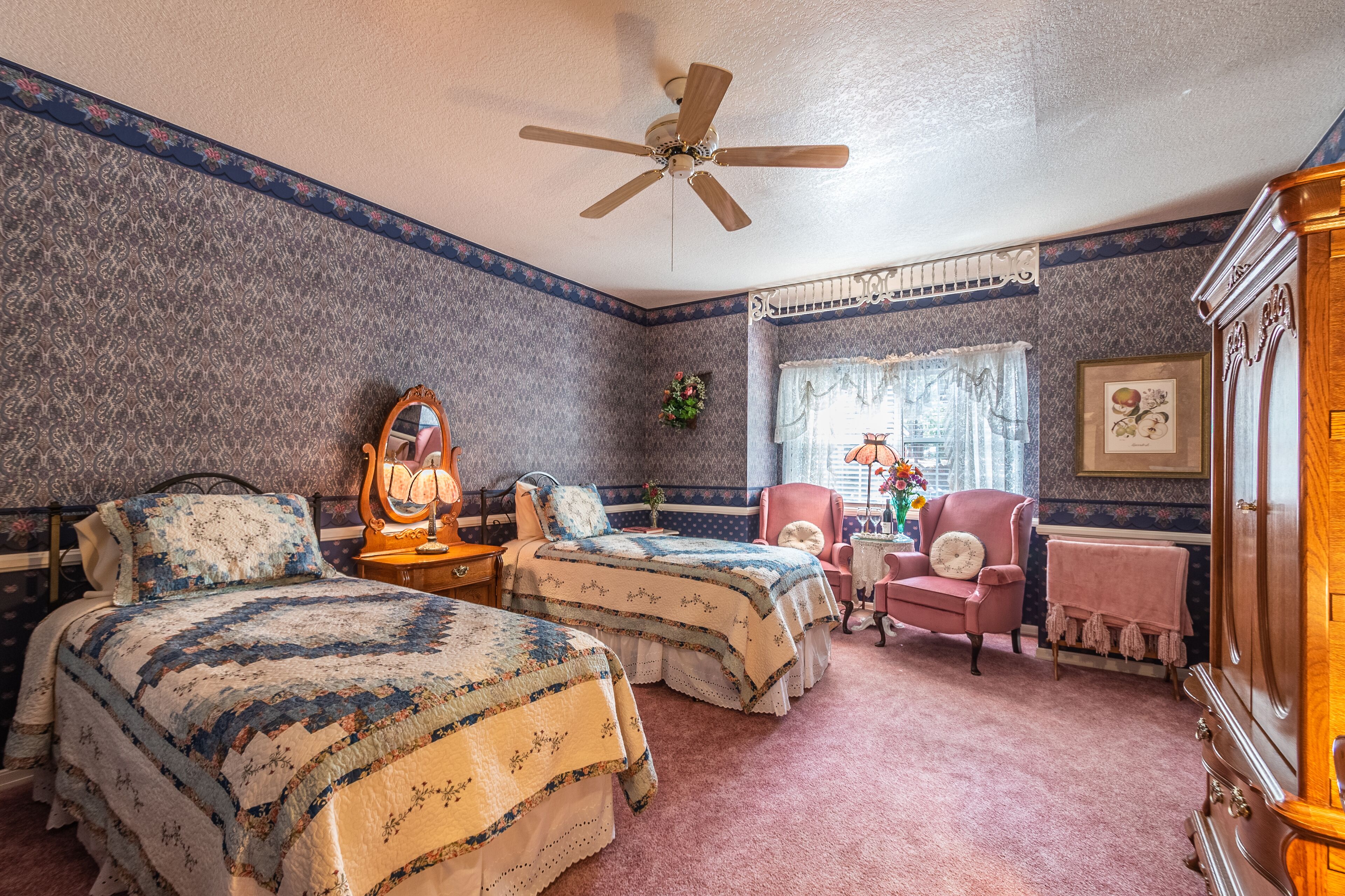 A bedroom with two beds, a ceiling fan, a dresser, a chair, and a window with curtains.