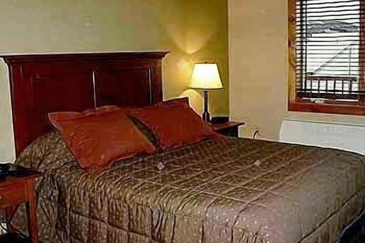 A bedroom with a wooden headboard, a single bed with a patterned bedspread, a bedside lamp, and a window with blinds.