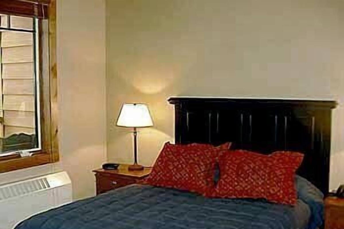A bedroom with a bed, a nightstand, a lamp, and a window with blinds.