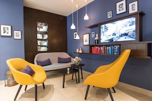 A living room with a sofa, two yellow chairs, a coffee table, and a television displaying a cityscape. - Hotel Stella Maris (Hamburg)