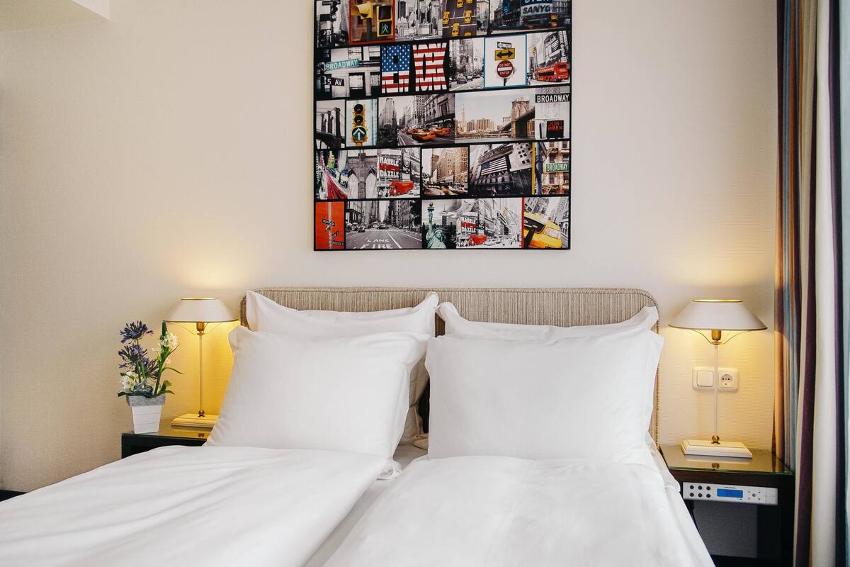 Habitación de hotel con una cama bien hecha, dos lámparas de noche, una obra de arte en la pared y una mesita con una lámpara y un arreglo floral.