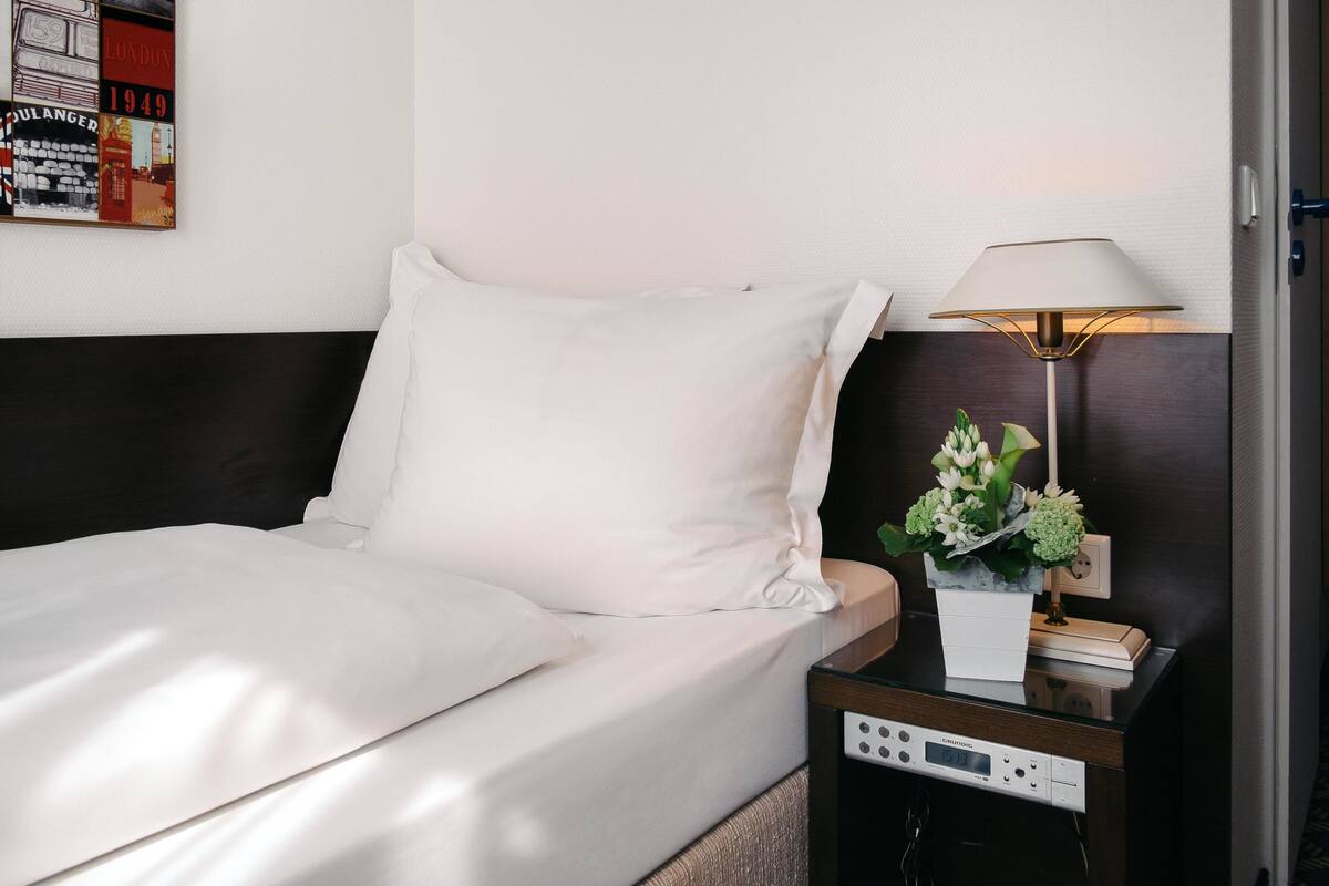 Una cama bien hecha con sábanas blancas, una mesita de noche con una lámpara, un reloj y una pequeña planta.