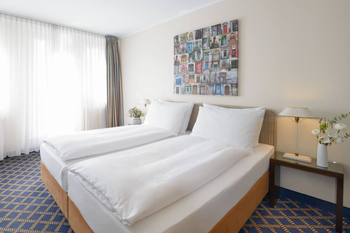 Habitación de hotel con dos camas, un ventanal con cortinas translúcidas, un cuadro colgado en la pared, una mesita de noche con lámpara y una alfombra estampada.