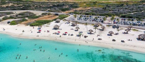Vue aérienne d’un complexe hôtelier en bord de mer, avec un parking, des parasols de plage et une mer turquoise d’une clarté exceptionnelle.