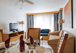 A hotel room with a dining table set for two, a television, a sofa, and a window with curtains.