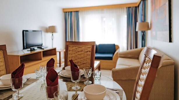 Ein Hotelzimmer mit Esstisch für vier Personen, einem Fernseher, einer Couch und einem großen Fenster mit Vorhängen.