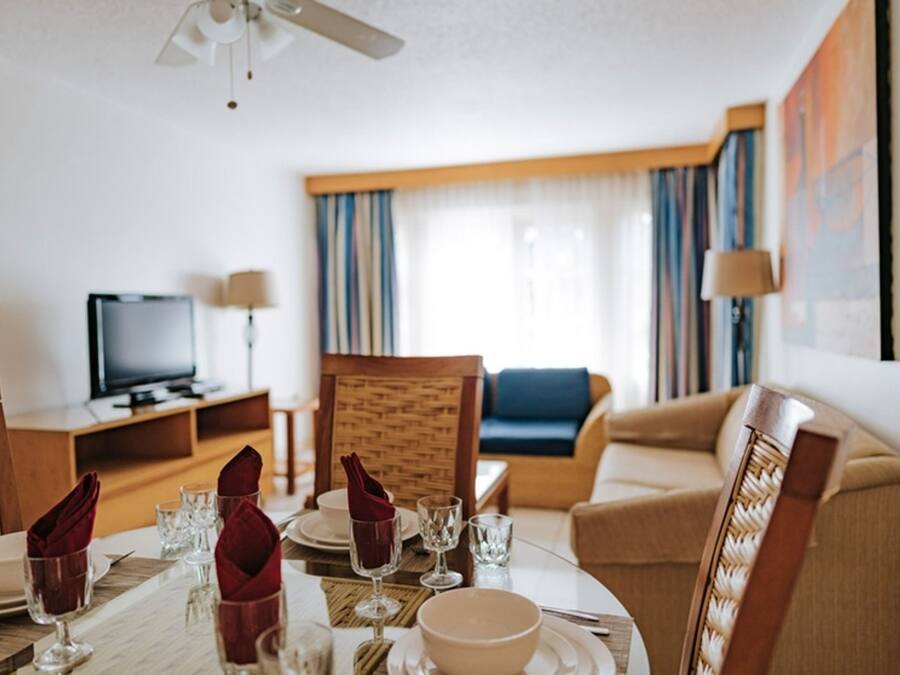 Ett hotellrum med ett matbord för fyra personer, en tv, en soffa och ett stort fönster med gardiner.