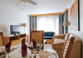 Habitación de hotel con una mesa de comedor para cuatro, televisor, sofá y un amplio ventanal con cortinas.