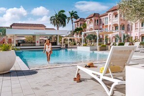 Zona de piscina con sillones de descanso, una mujer caminando y un edificio con tejado de azulejos al fondo.