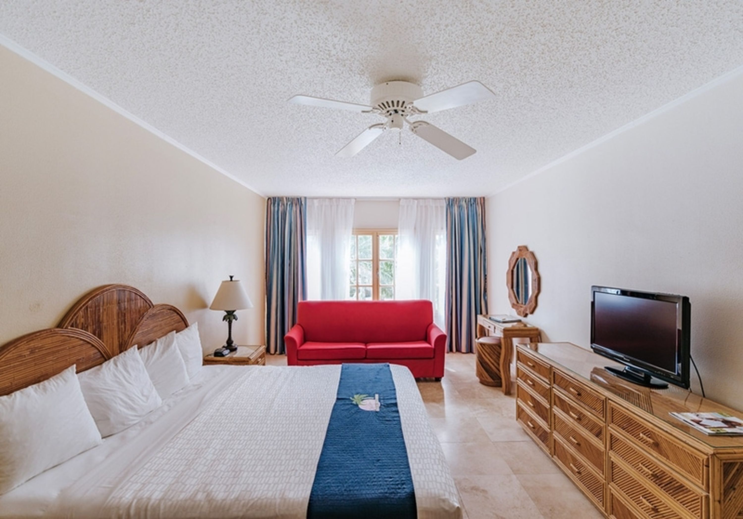 A hotel room with a bed, a red sofa, a television, and a ceiling fan.