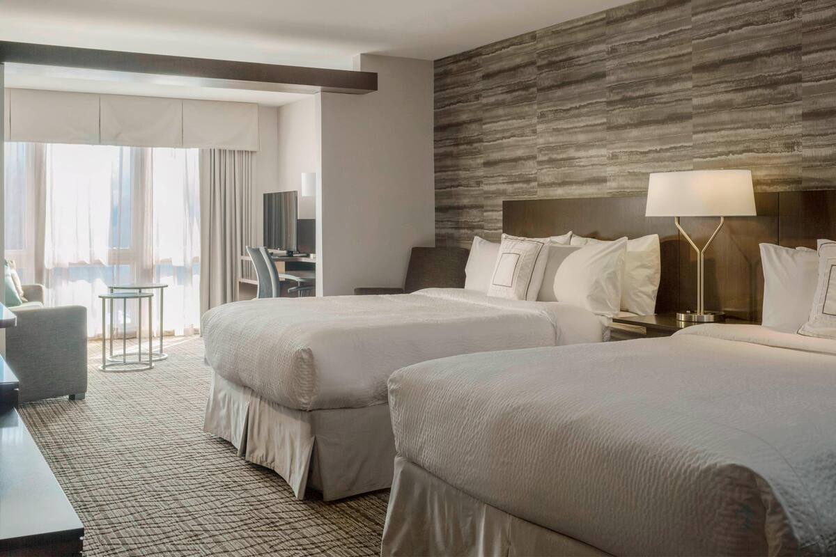 A hotel room with two beds, a desk, a chair, a TV, and a view of the outdoors.