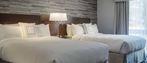 A hotel room with two beds, a modern headboard, a bedside lamp, and a large window with curtains.