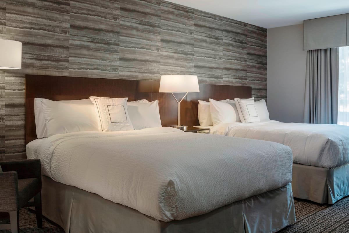 A hotel room with two beds, a wooden accent wall, a nightstand with a lamp, and a view of the city through large windows.