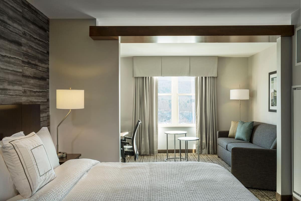 A hotel room with a large bed, a desk with a chair, a sofa, and a window with curtains.