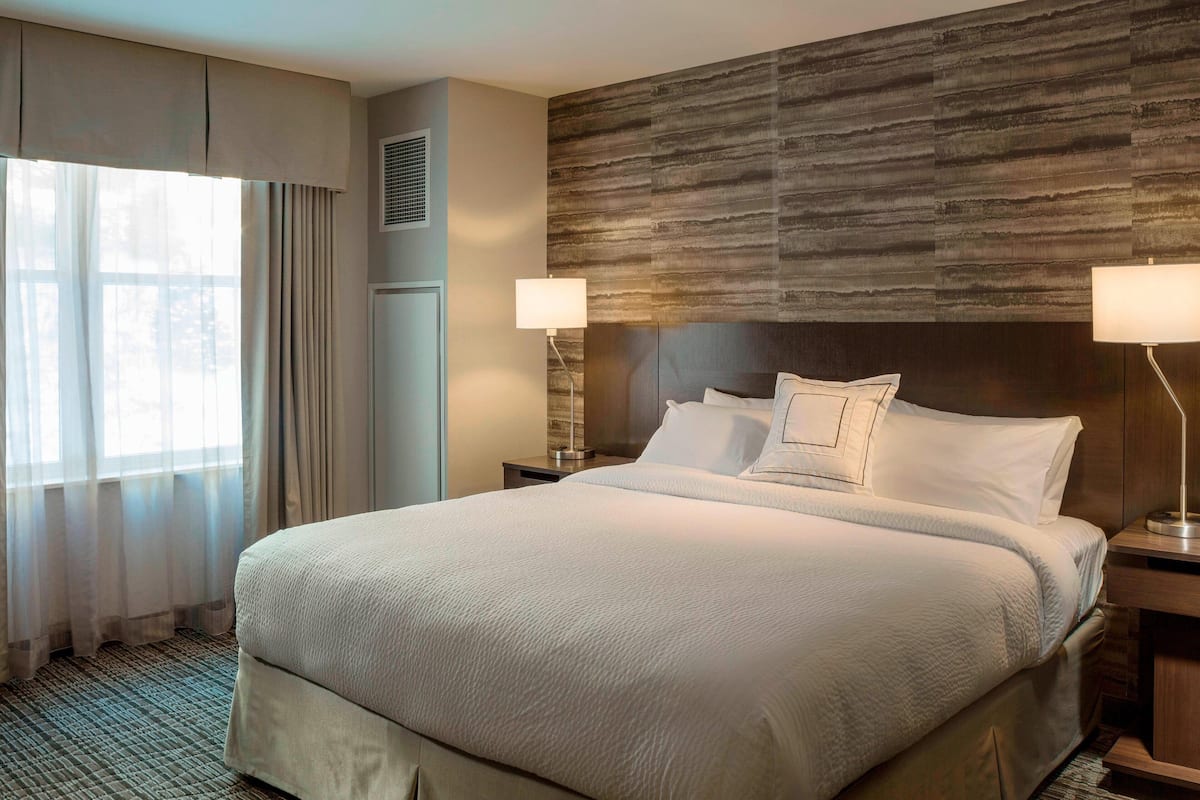 A hotel room with a large bed, two bedside lamps, a wooden headboard, and a window with curtains.