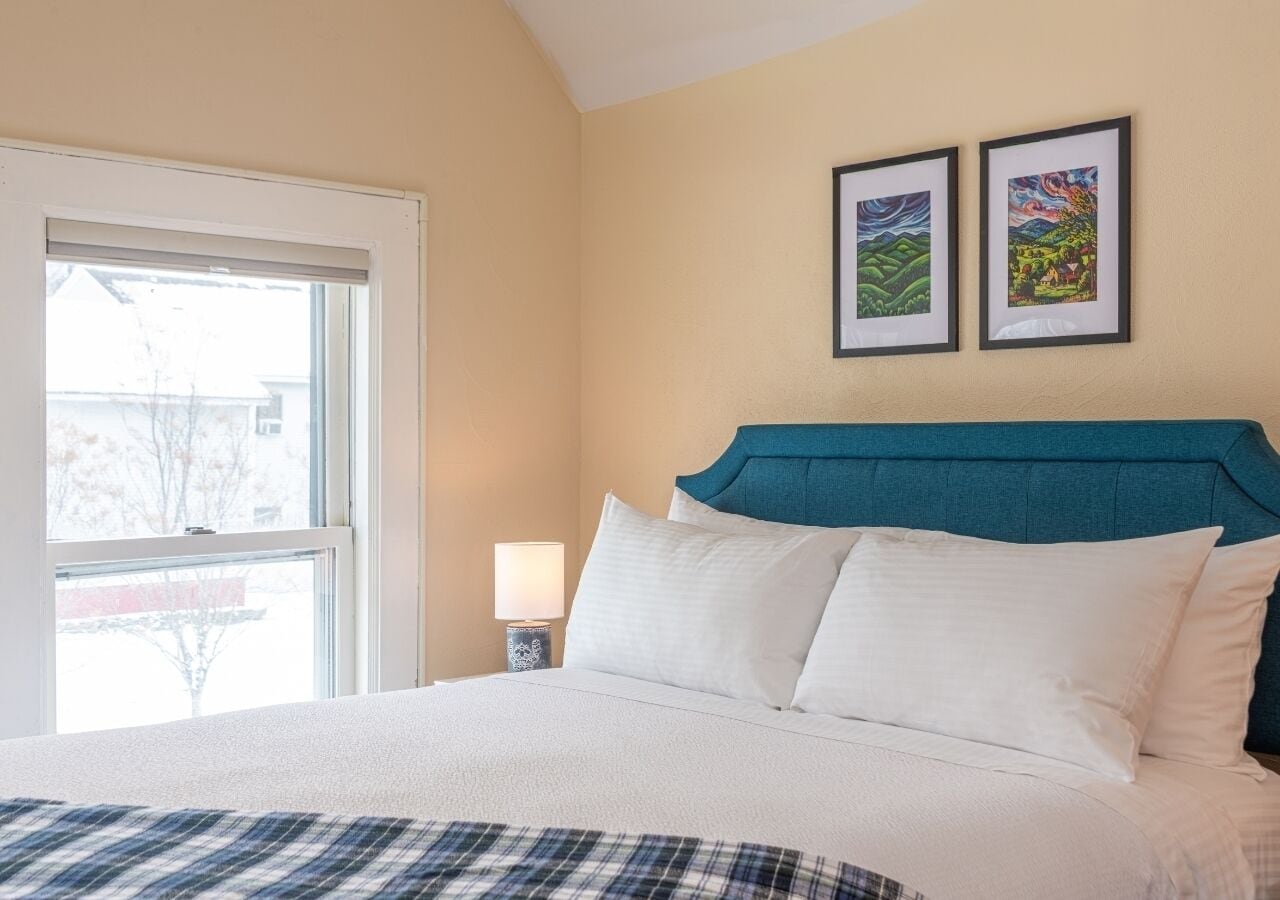 A hotel room with a bed, a nightstand, a window, and two framed pictures on the wall.