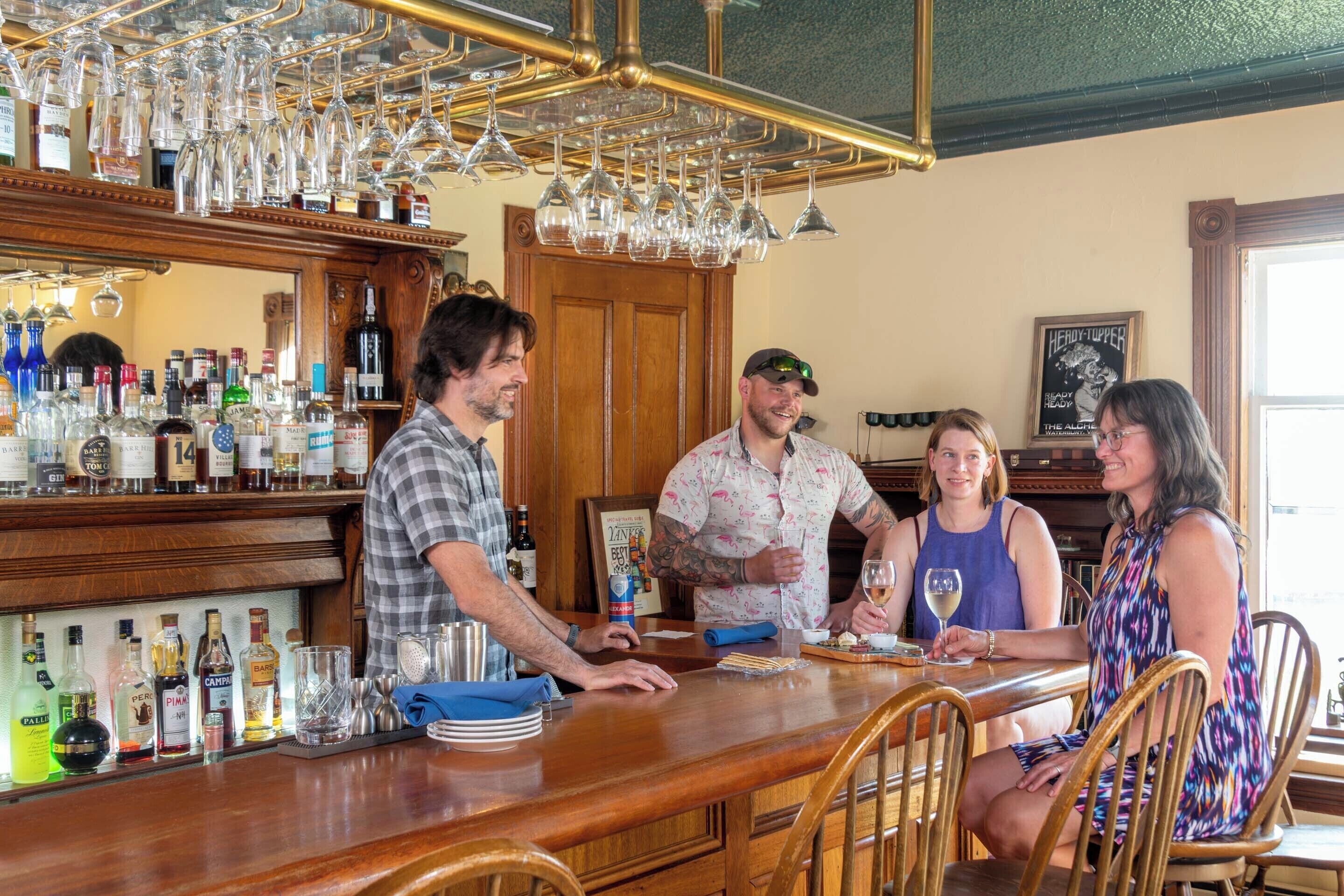 A bar area with a wooden counter, various bottles, and glasses. There are chairs around the bar and a framed picture on the wall.