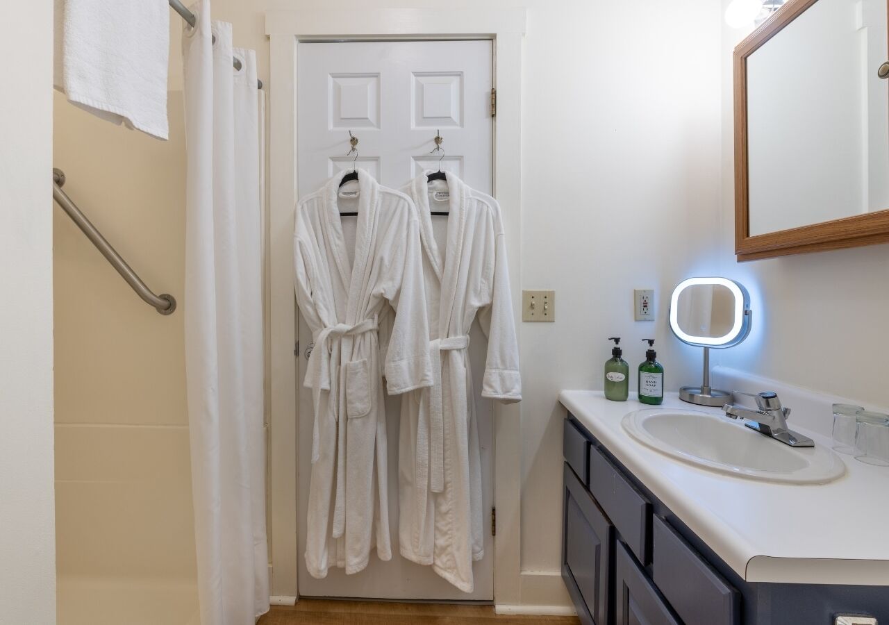 Un baño con un lavamanos blanco, un espejo y dos albornoces blancos colgados de una puerta.