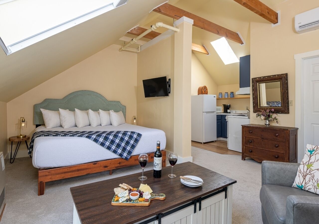 A bedroom with a bed, a breakfast table with cheese and wine, a kitchenette, and a mirror.
