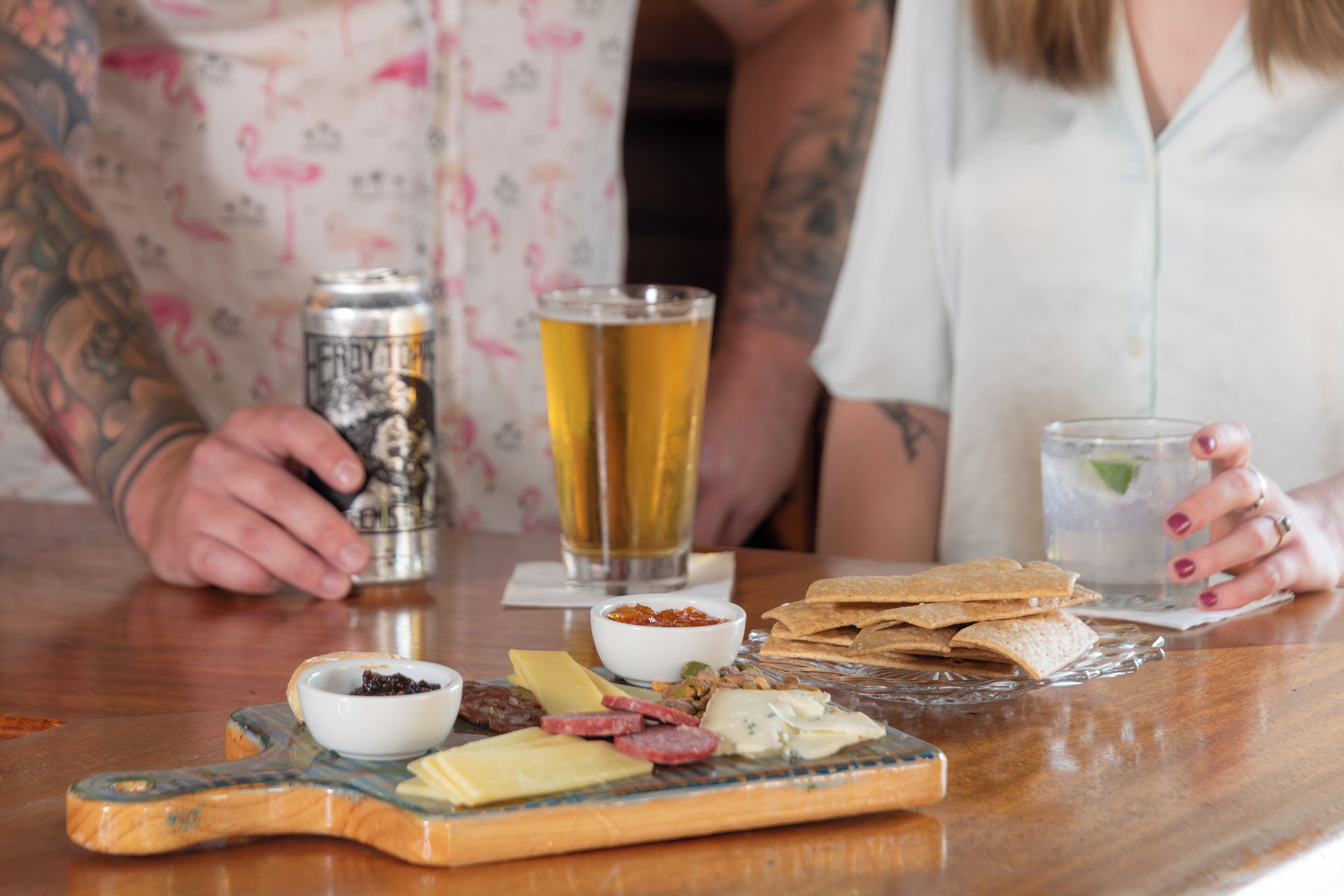 A wooden table with a variety of cheeses, crackers, and a can of beer.