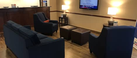 A hotel lobby with a reception desk, seating area with sofas and chairs, and a television displaying information about services.