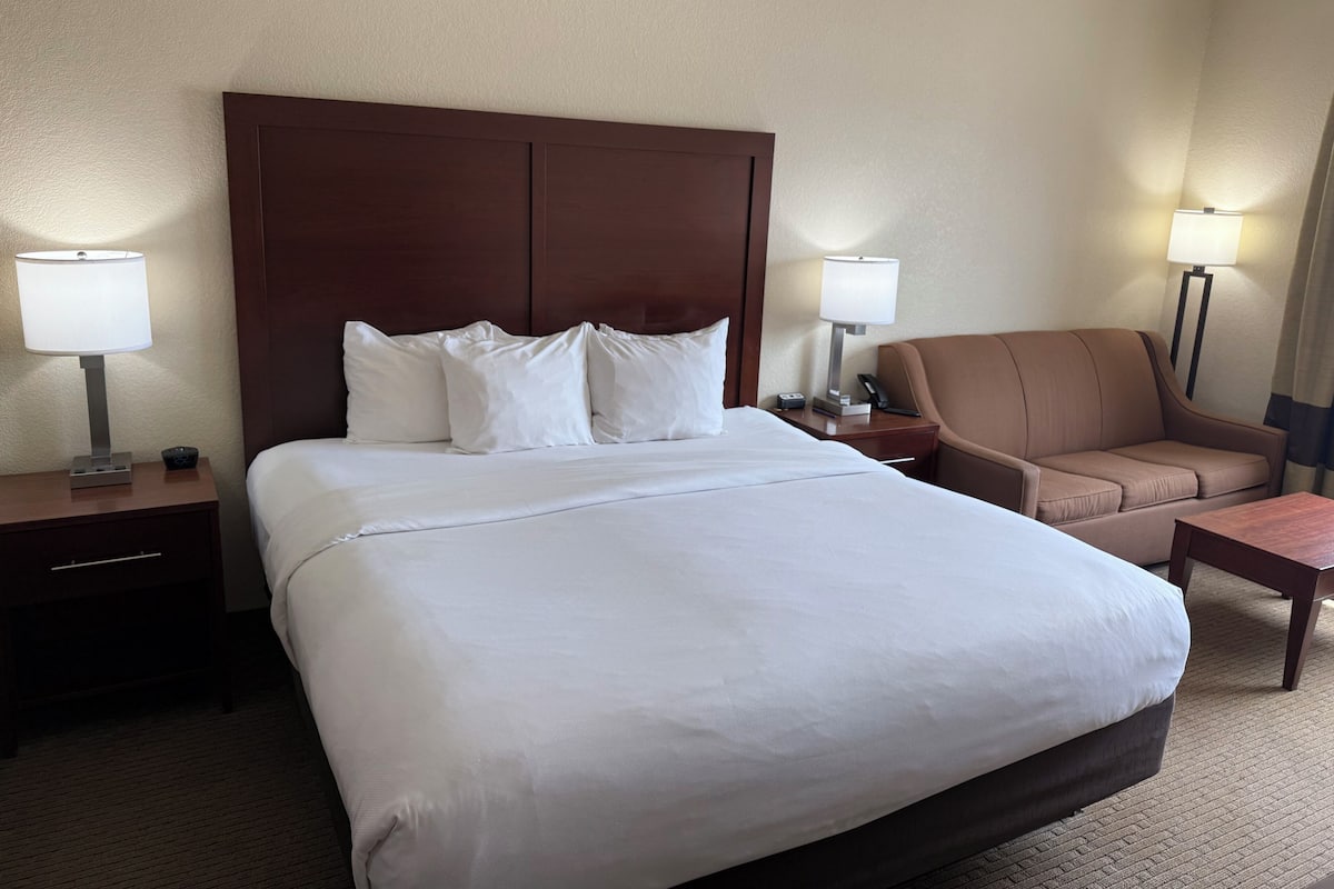 Una habitación de hotel con una cama grande, un cabecero marrón, dos mesitas de noche con lámparas, un sillón marrón y una pequeña mesa de madera.