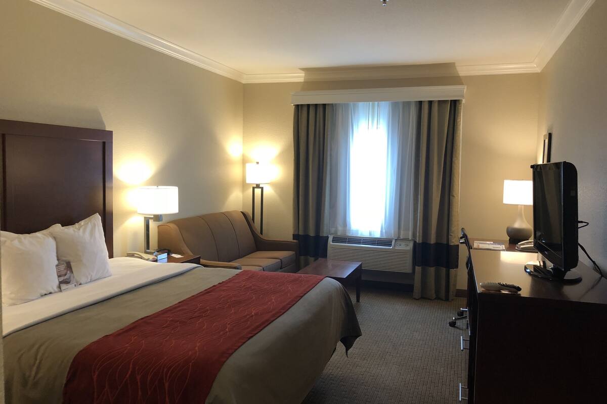 Una habitación de hotel con cama, sofá, escritorio, televisor y ventana con cortinas.