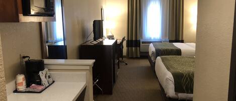 A hotel room with a kitchenette, a desk, a TV, two beds, and a window with curtains.
