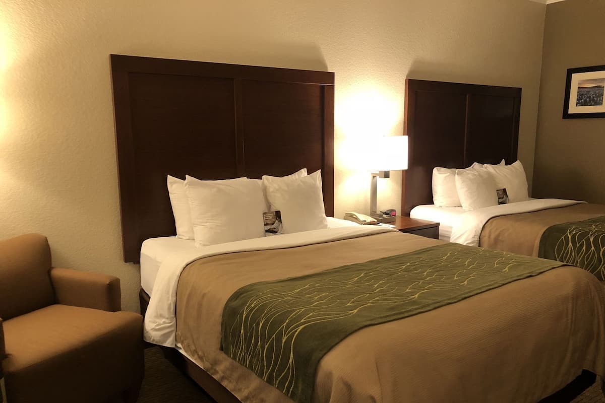 Habitación de hotel con dos camas, una silla y una mesita de noche con lámpara.