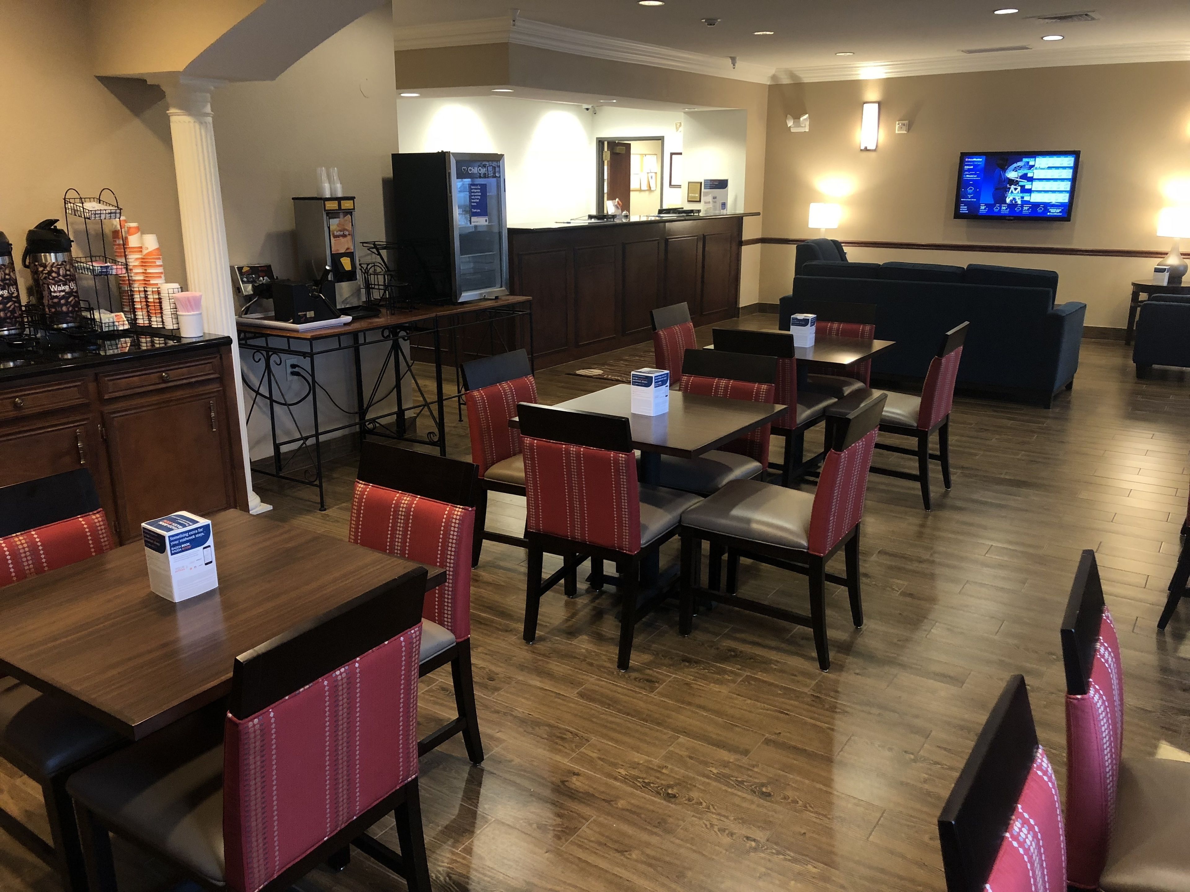 A hotel dining area with wooden tables and red upholstered chairs, a kitchen area, and a TV mounted on the wall.