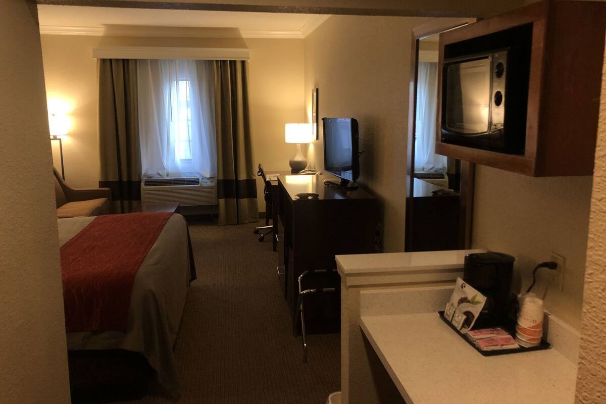 Una habitación de hotel con cama, escritorio con computadora, televisor, espejo y ventana con cortinas.