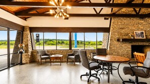 A spacious room with a stone fireplace, large windows offering a view of the outdoors, and a ceiling with exposed beams. - Segovia Lodge Junction TX I -10 (Junction)