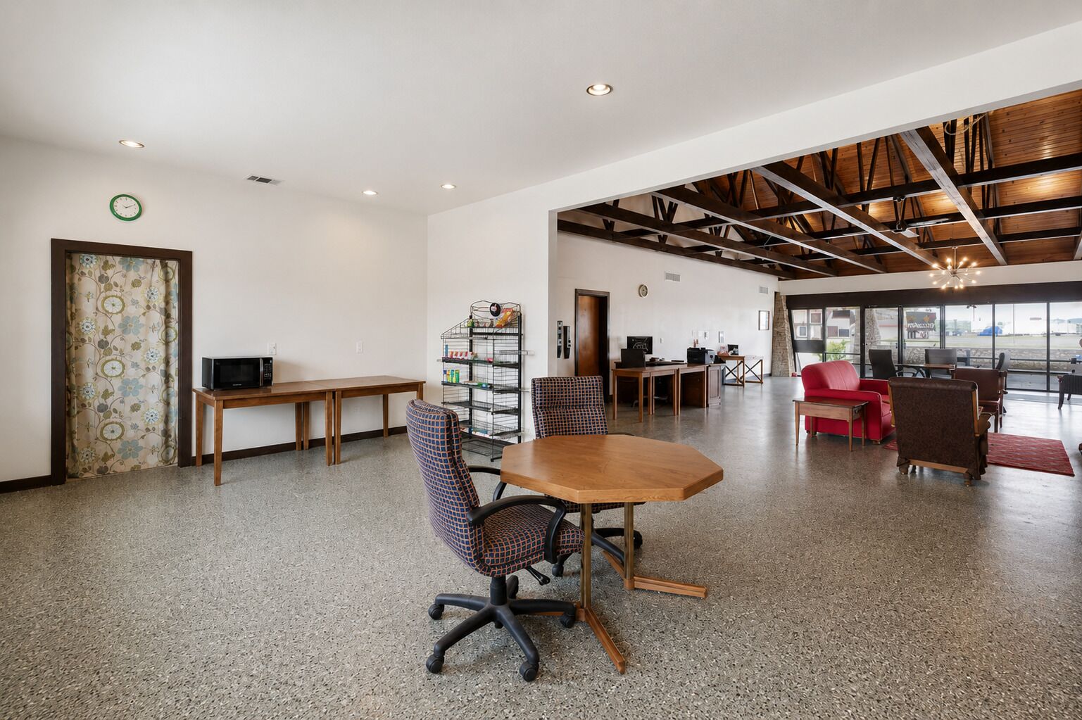 A spacious room with a large table, chairs, a red couch, and a kitchenette area.