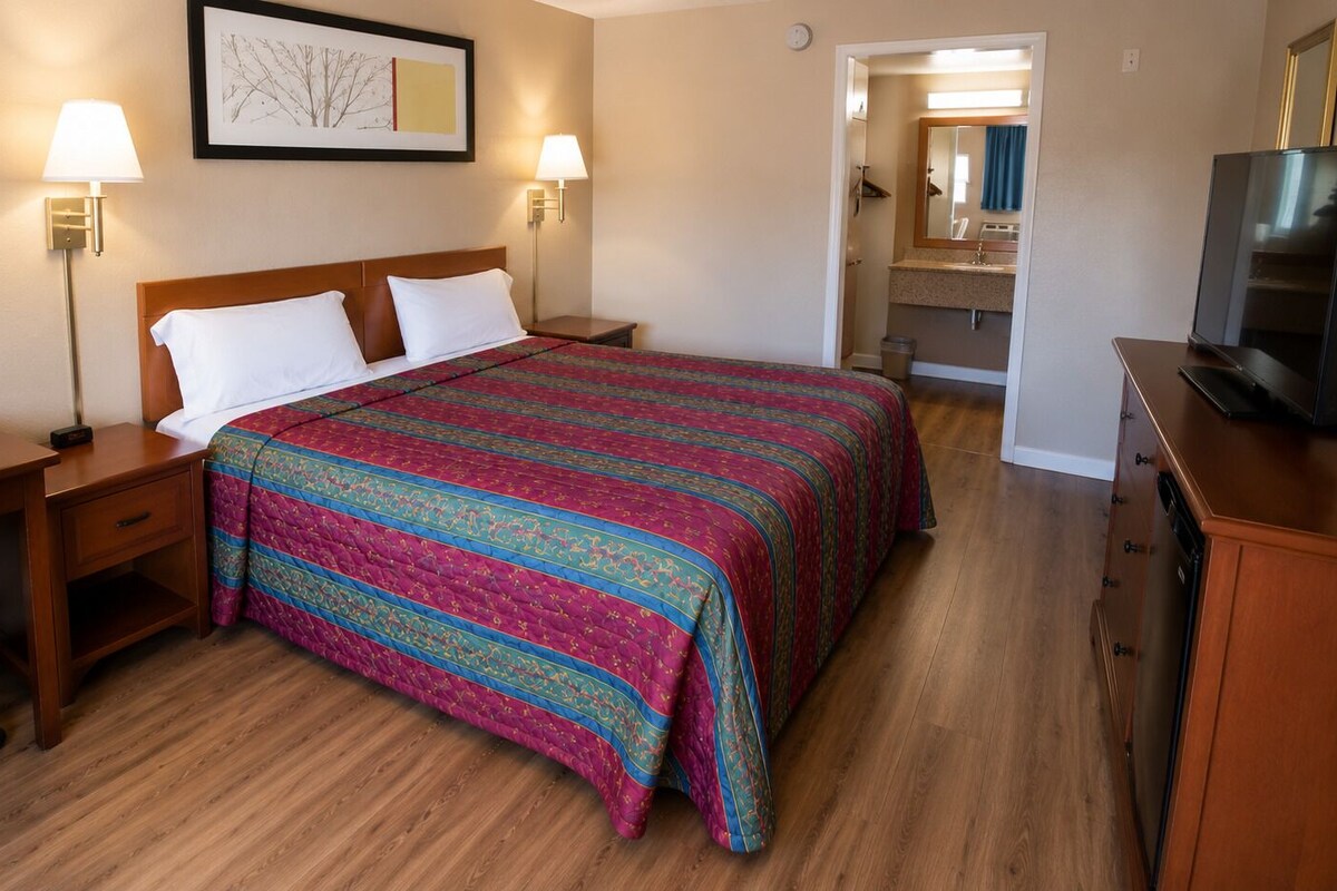 A hotel room with a bed, bedside tables, a desk, a chair, a television, and a bathroom visible through an open door.