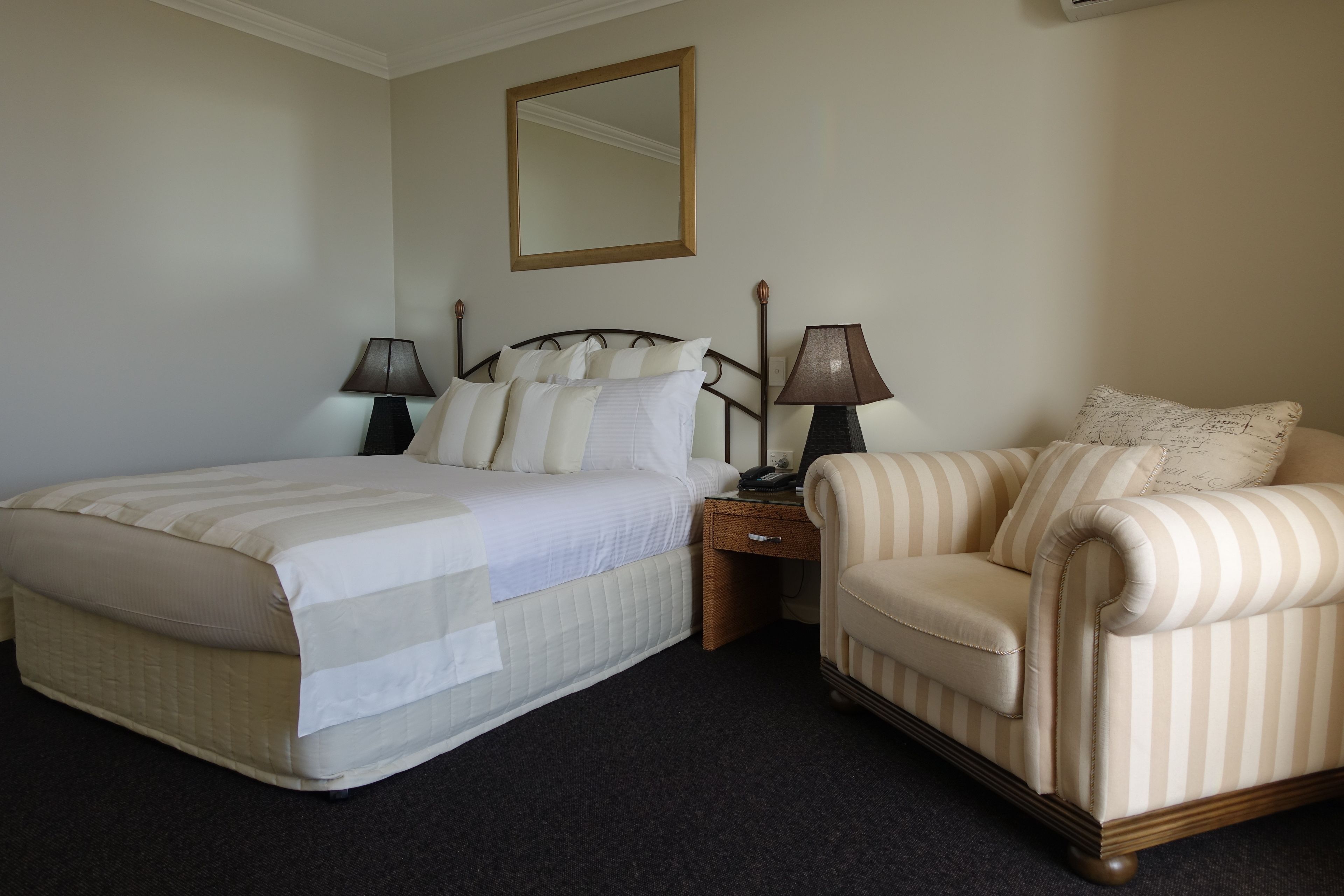 Una camera d'albergo con un letto, un comodino, una lampada, una sedia e uno specchio a parete.