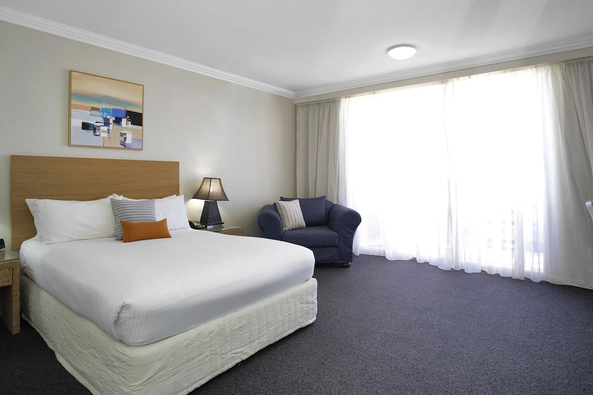 A hotel room with a bed, a nightstand, a lamp, a chair, and a large window with curtains.