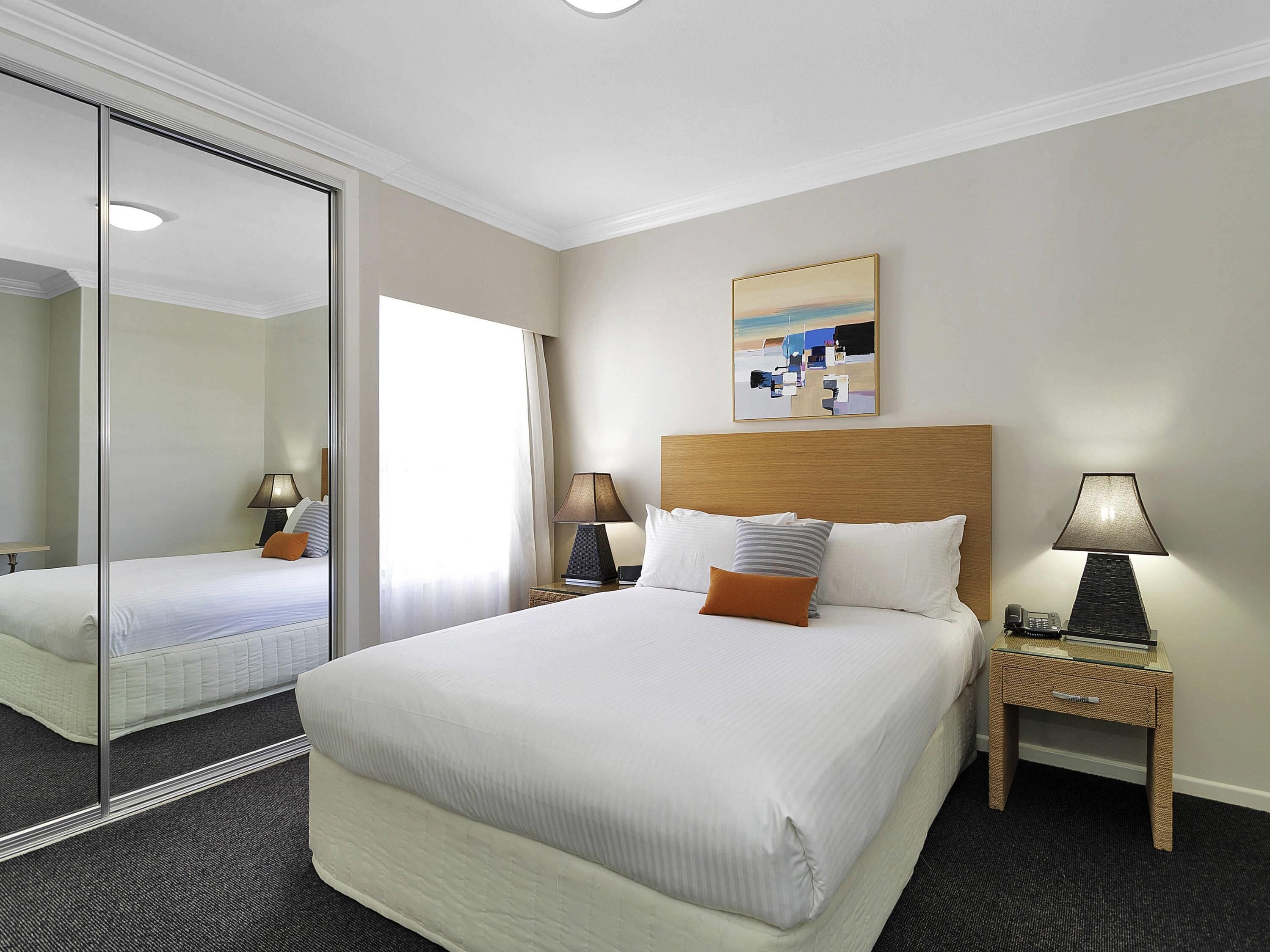 A hotel room with a large bed, a bedside table with a lamp, a bedside phone, a mirror, and a painting on the wall.