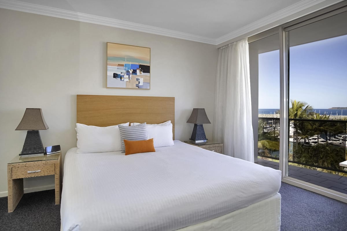 A hotel room with a large bed, two bedside tables with lamps, a painting on the wall, and a view of the sea through a large window.
