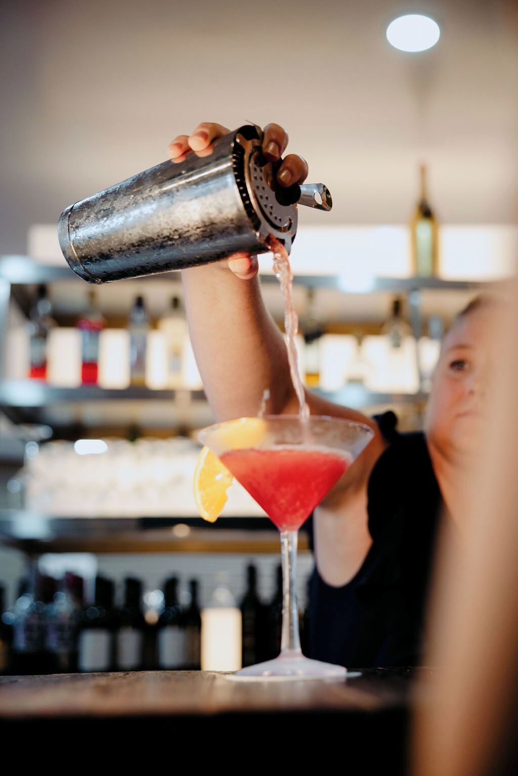 A person pouring a drink into a martini glass with an orange garnish.