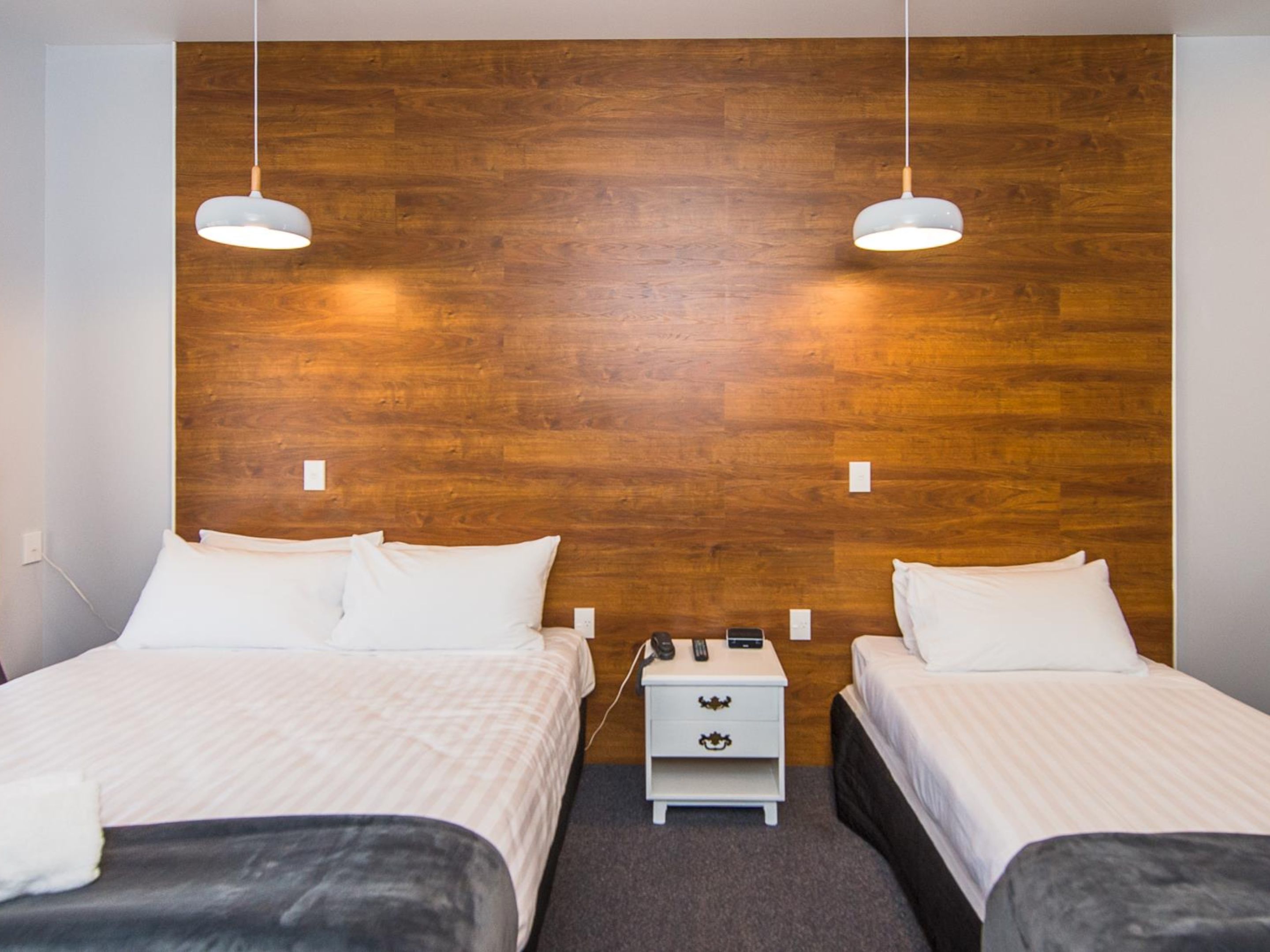 Habitación de hotel con dos camas, cabecero de madera, ropa de cama blanca, mesitas de noche con lámparas y suelo cubierto de alfombra.