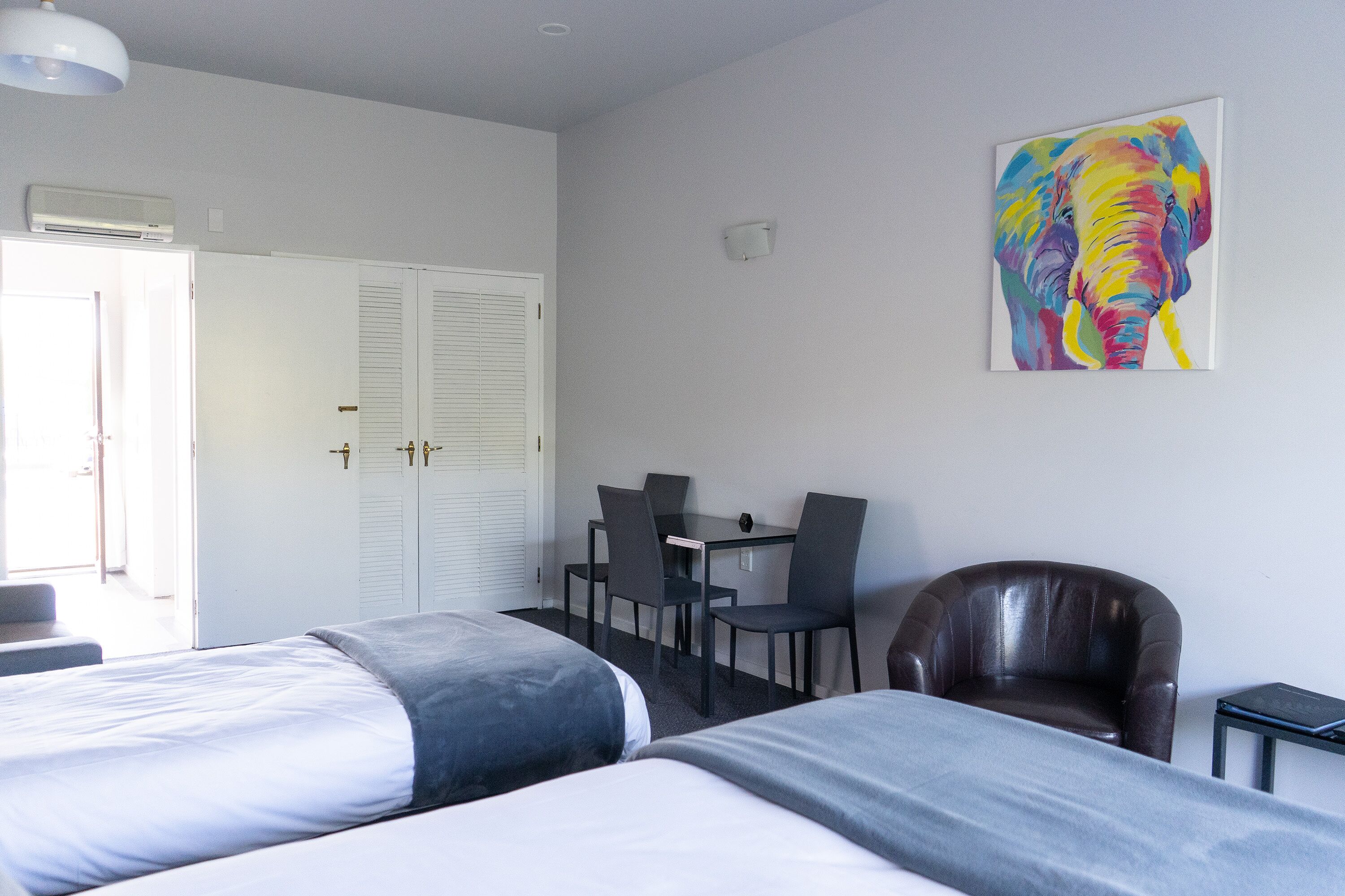 Habitación de hotel con dos camas, un escritorio con sillas, un sillón de cuero y un colorido cuadro de un elefante en la pared.